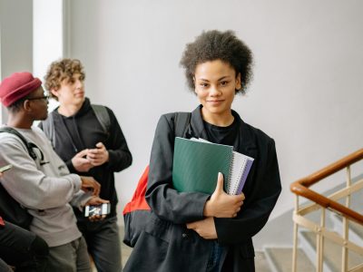 Young woman with afro hair holding notebooks, standing in university hallway, smiling confidently.